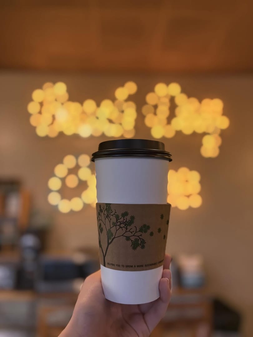 Hand holding a white coffee cup with a tree-patterned sleeve against blurred golden lights.