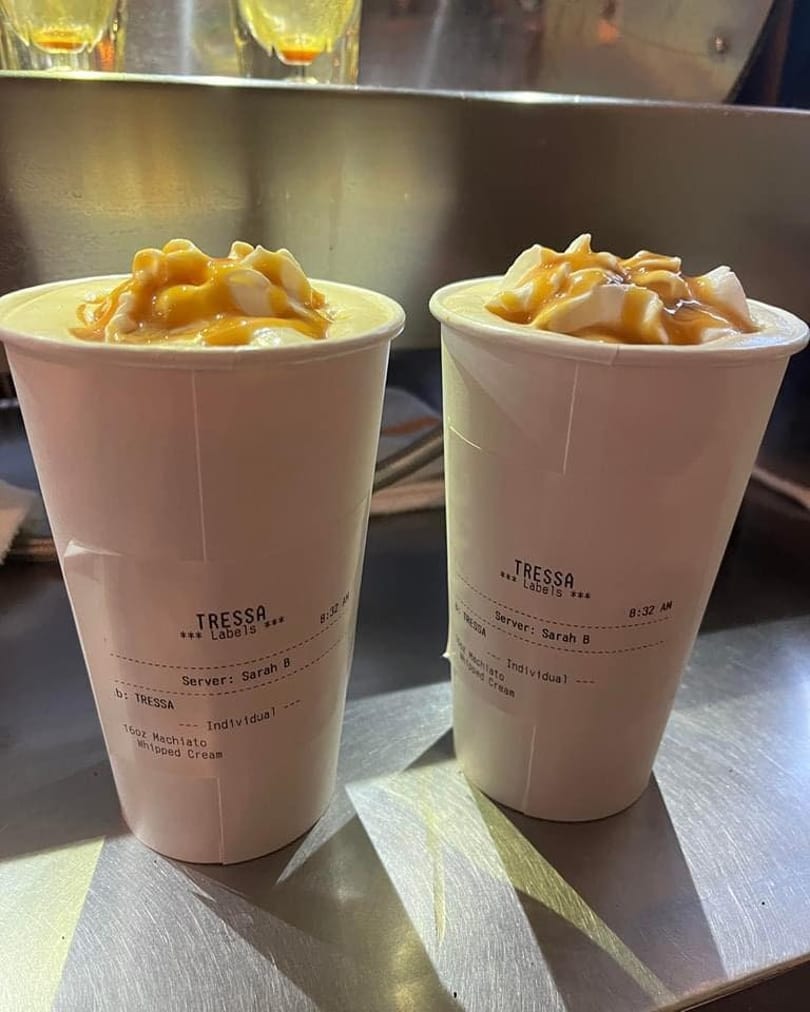 Two Tressa Labels macchiatos with whipped cream and caramel drizzle on a metal counter.