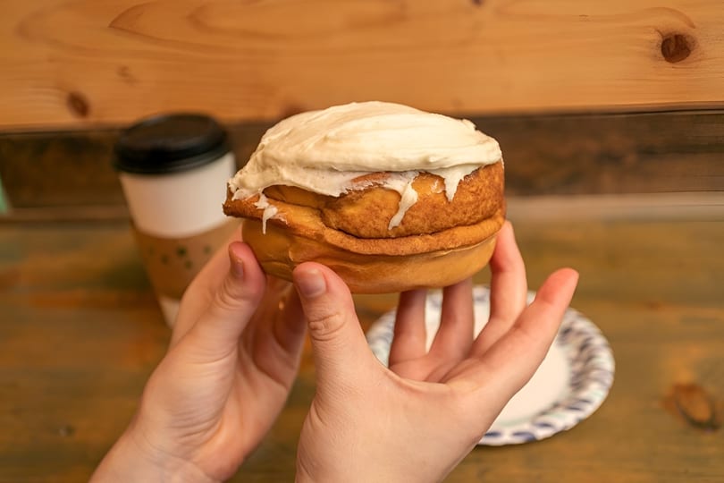Hands holding a large, frosted cinnamon roll with a coffee cup and paper plate in the background.
