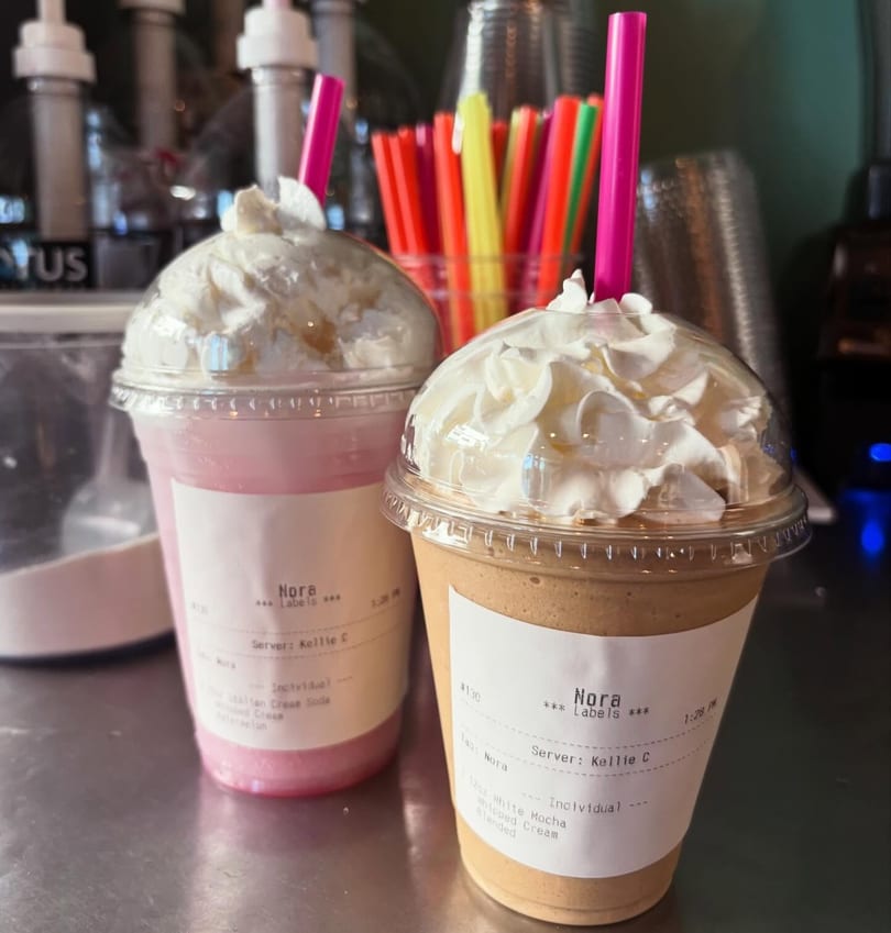 Two Nora brand blended drinks with whipped cream and pink straws on a counter.