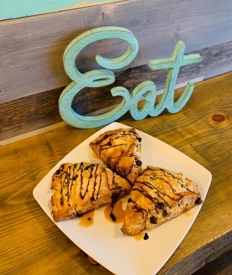 Three chocolate chip scones drizzled with caramel and chocolate on a white plate next to a blue "Eat" sign.