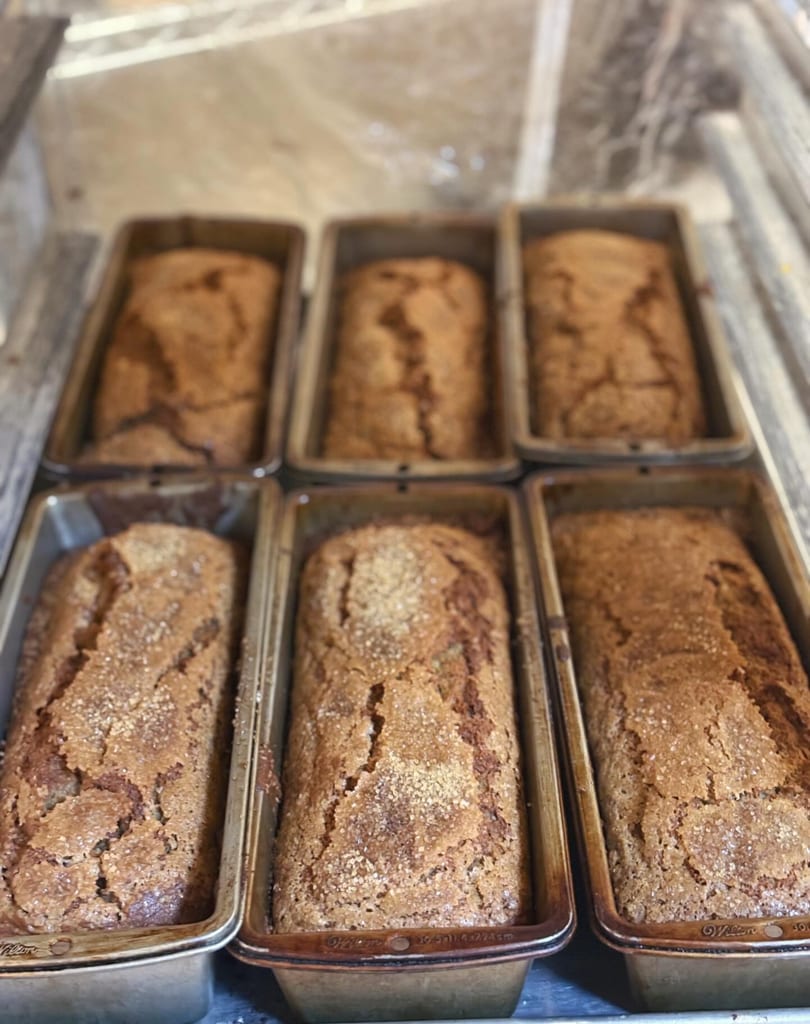 Six freshly baked loaves of banana bread in metal pans, topped with sparkling sugar.
