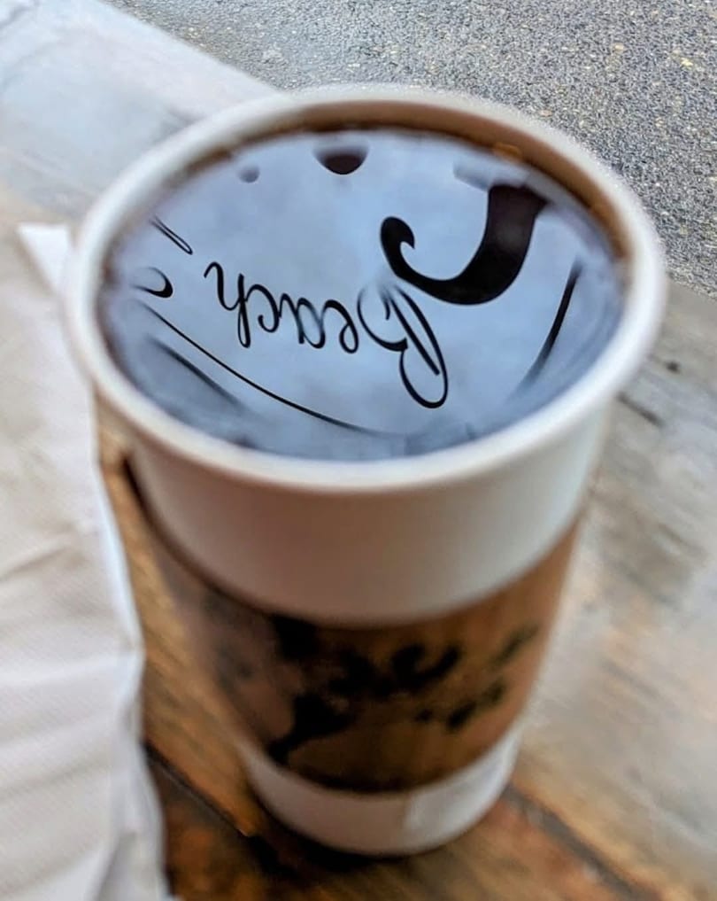 Close-up of a coffee cup with "Beach" reflected on the dark surface of the drink.