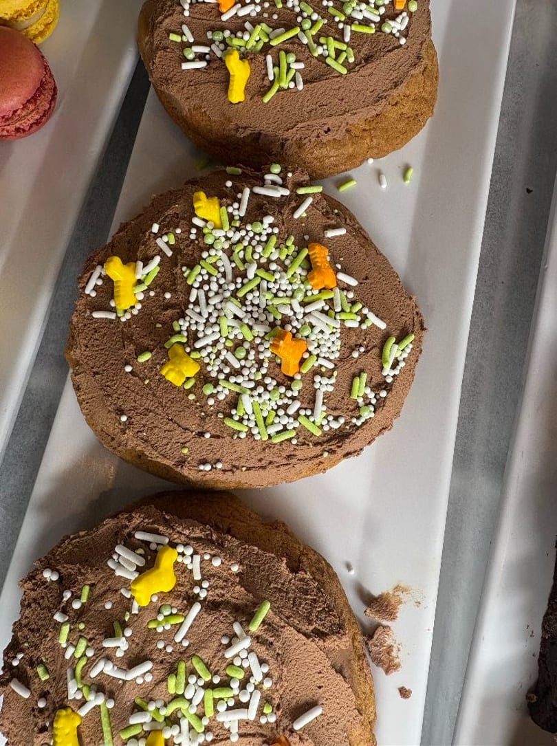 Three chocolate frosted cookies with green, white, yellow, and orange sprinkles on a white tray.